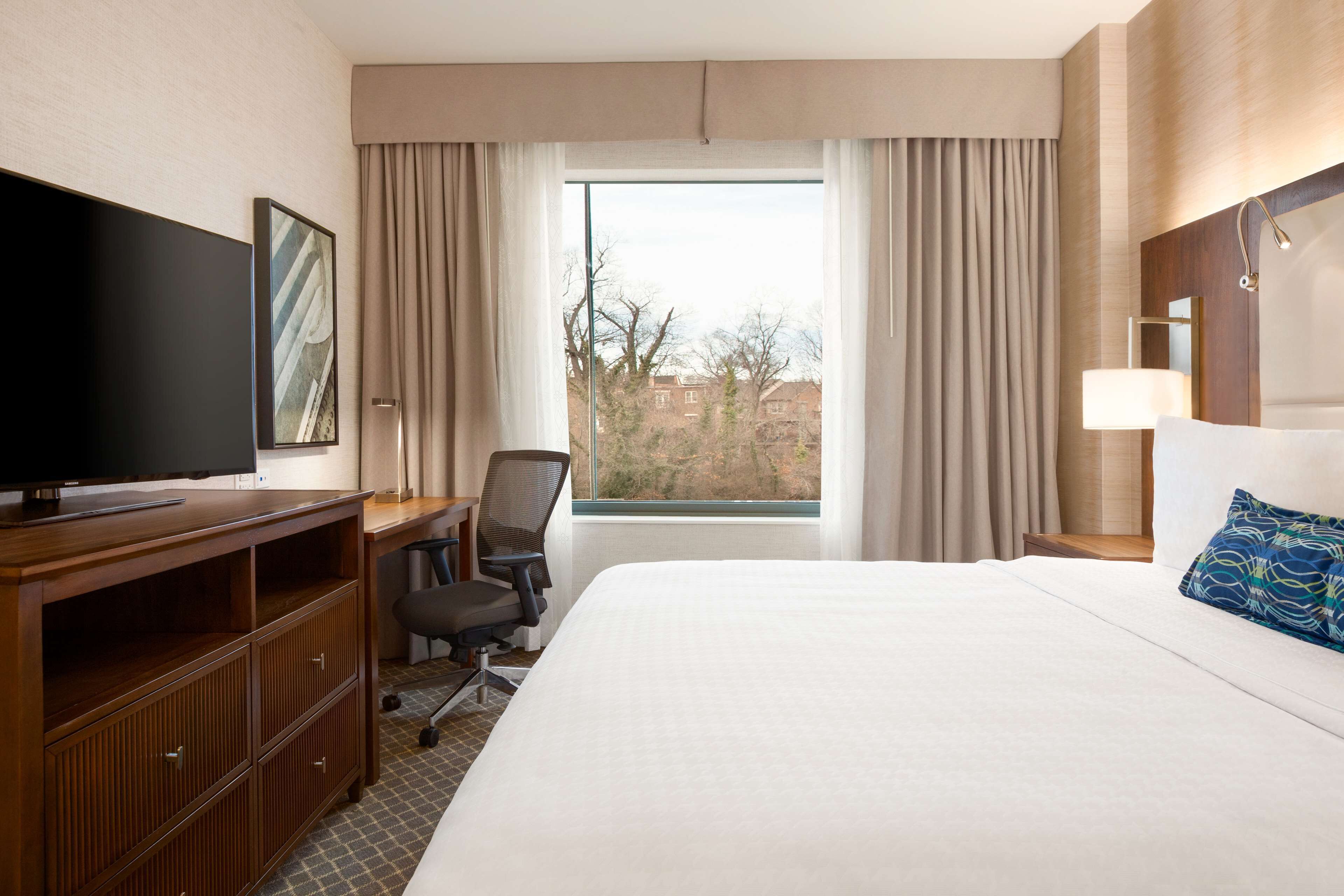 A hotel room with a large bed, a desk with a television, a chair, and a window with curtains.