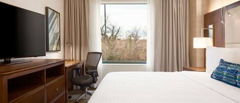 A hotel room with a large bed, a desk with a television, a chair, and a window with curtains.