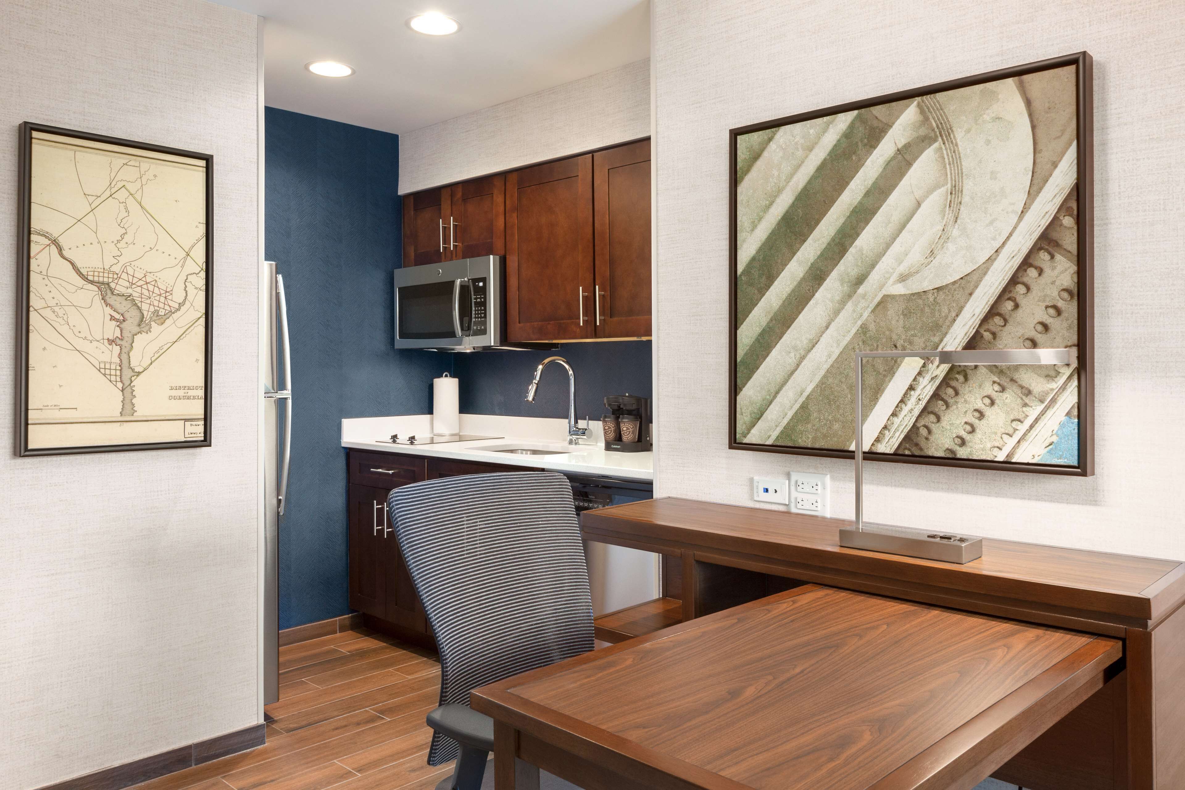 A compact kitchen with a microwave, sink, and cabinets. There is a wooden desk and a chair in the room.