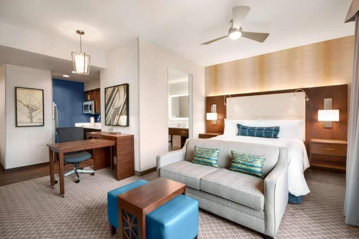 Une chambre d’hôtel équipée d’un lit, d’un bureau, d’une chaise, d’un canapé, d’un ventilateur de plafond et d’un téléviseur fixé au mur.