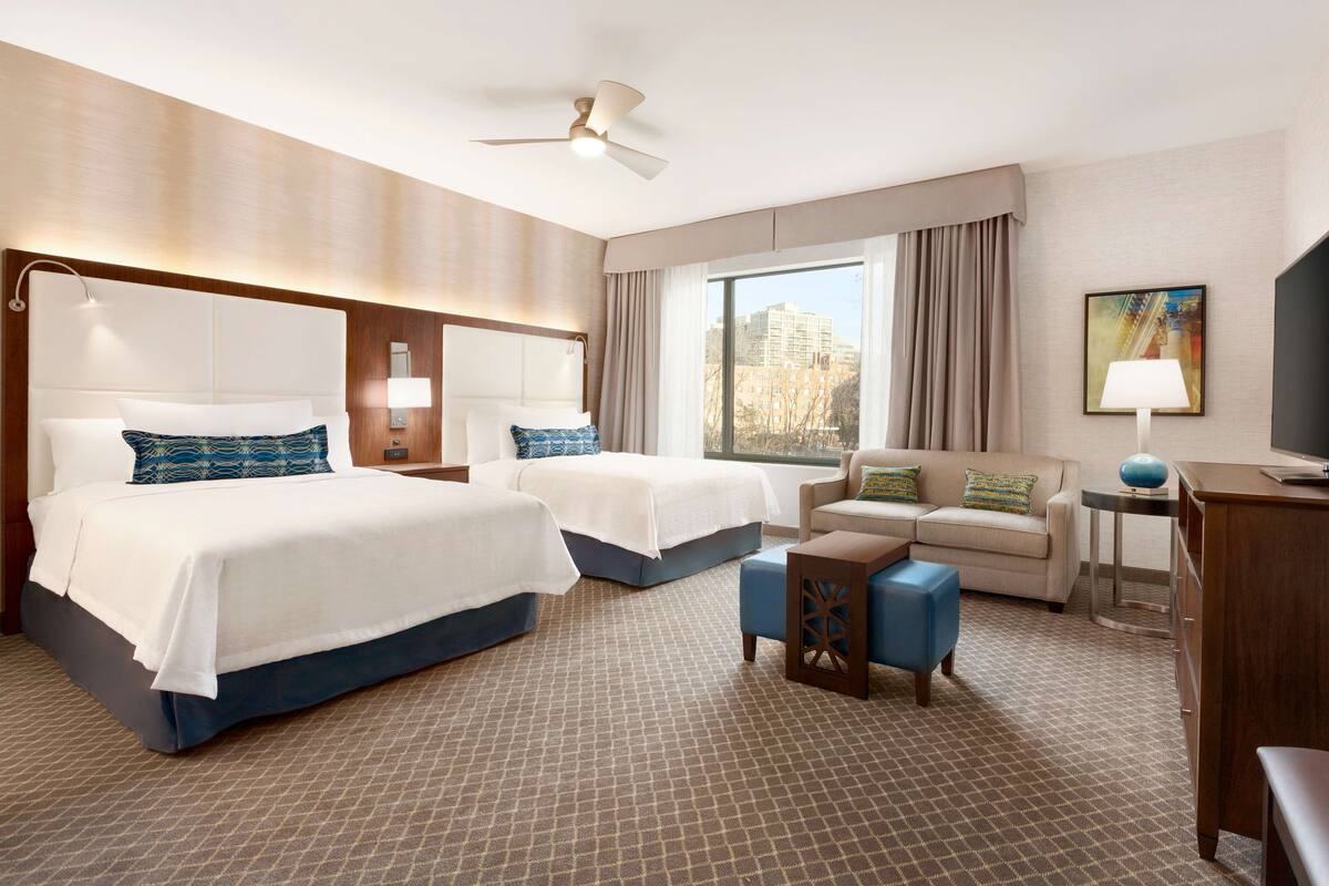 Une chambre d’hôtel avec deux lits, un canapé, un bureau et une grande fenêtre donnant sur la ville.