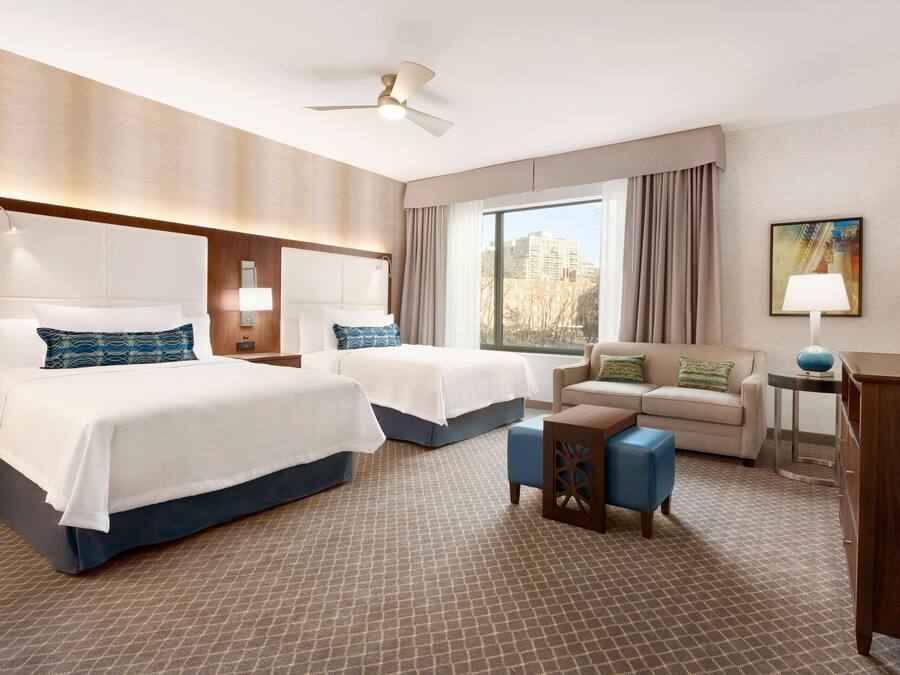 A hotel room with two beds, a sofa, a desk, and a large window with a city view.