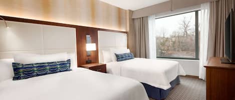 A hotel room with two beds, a ceiling fan, a large window with curtains, and a television on a wooden stand.