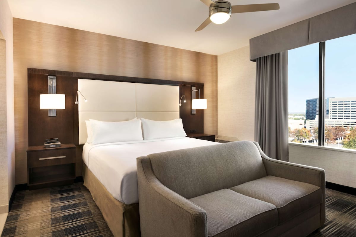 Une chambre d’hôtel avec un grand lit, une table de chevet, une lampe de lecture, un ventilateur de plafond et une vue sur la ville par la fenêtre.