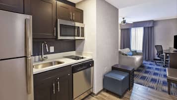 Quarto de hotel compacto com cozinha compacta, incluindo frigorífico, micro-ondas e máquina de lavar louça. Possui área de estar com sofá e uma pequena mesa, e uma televisão sobre um móvel.