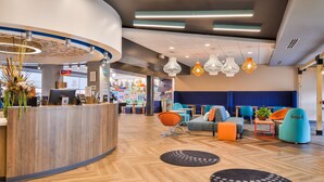 A modern reception area with a wooden desk, seating area with colorful chairs, and pendant lighting. - Tru by Hilton Stuttgart, AR (Stuttgart)