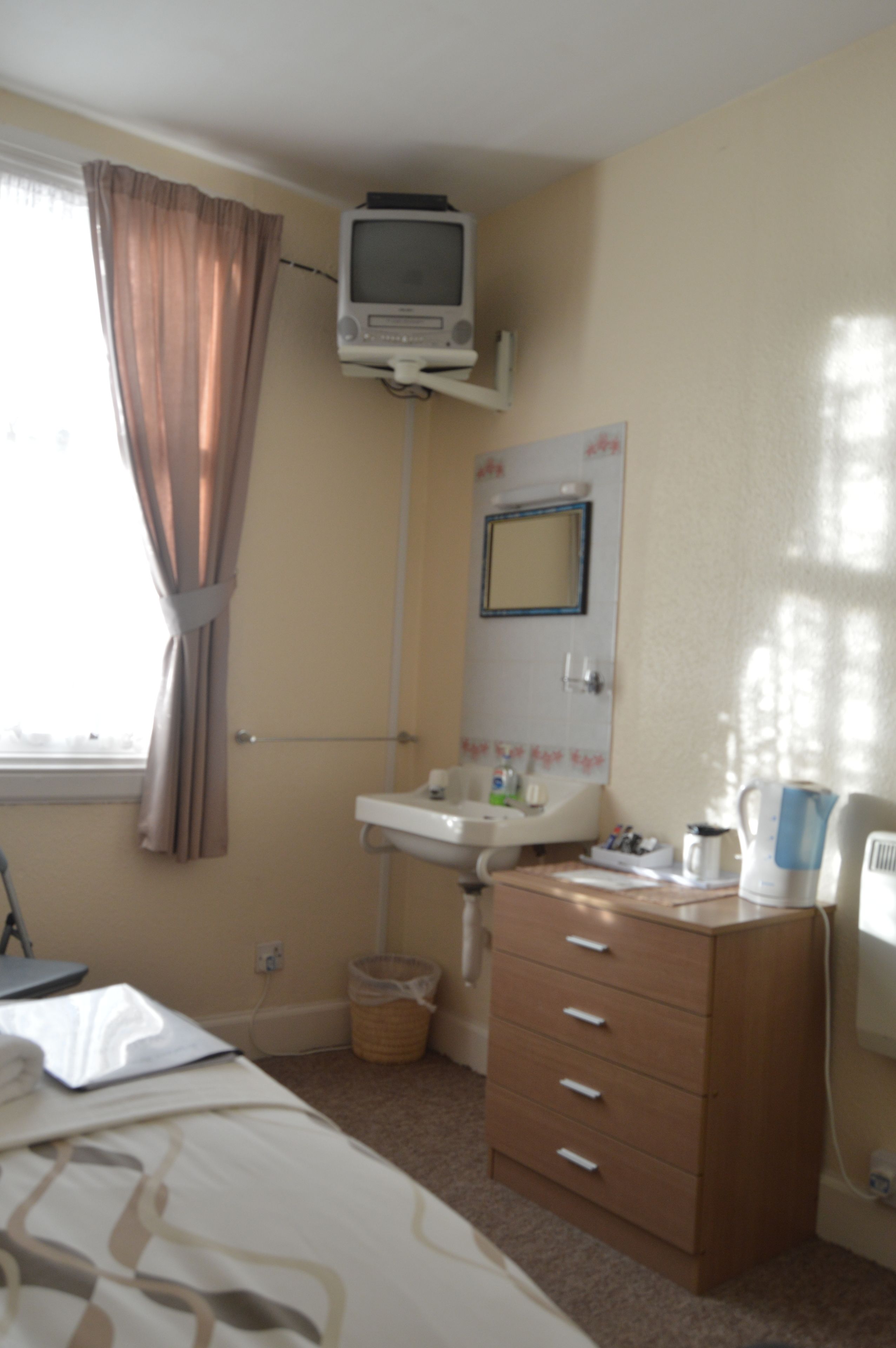 Habitación de hotel con cama, mesita de noche, cómoda, televisor montado en la pared, ventana con cortinas, lavamanos y espejo.