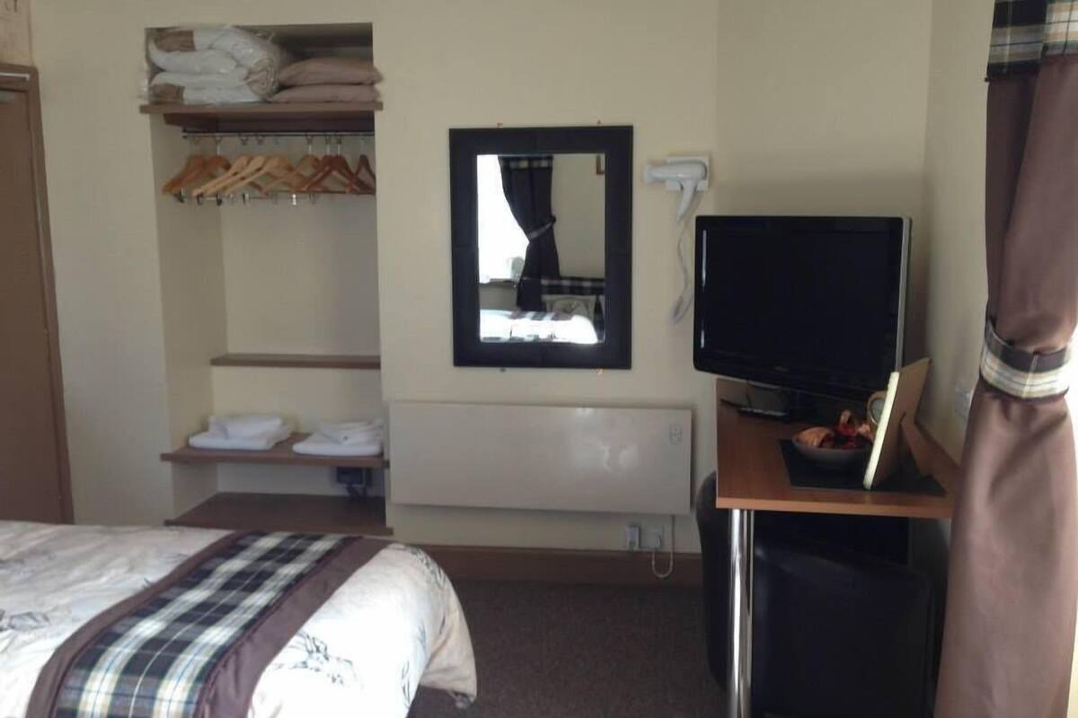 A hotel room with a bed, a TV, a mirror, a shelf with folded towels, and a coat rack.