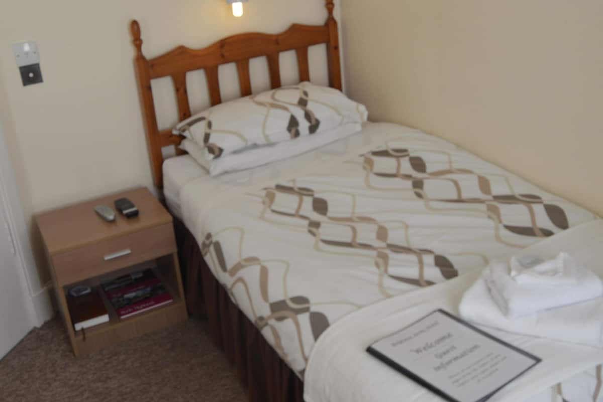 A single bed with a wooden headboard, a bedside table with a remote control, and a small shelf with books.