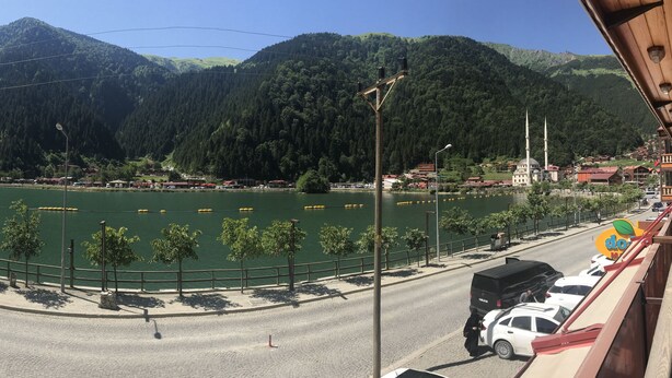 A view of a river with mountains in the background, a road with parked cars, and a building with a dome and minarets. - Doğa Motel (Çaykara)