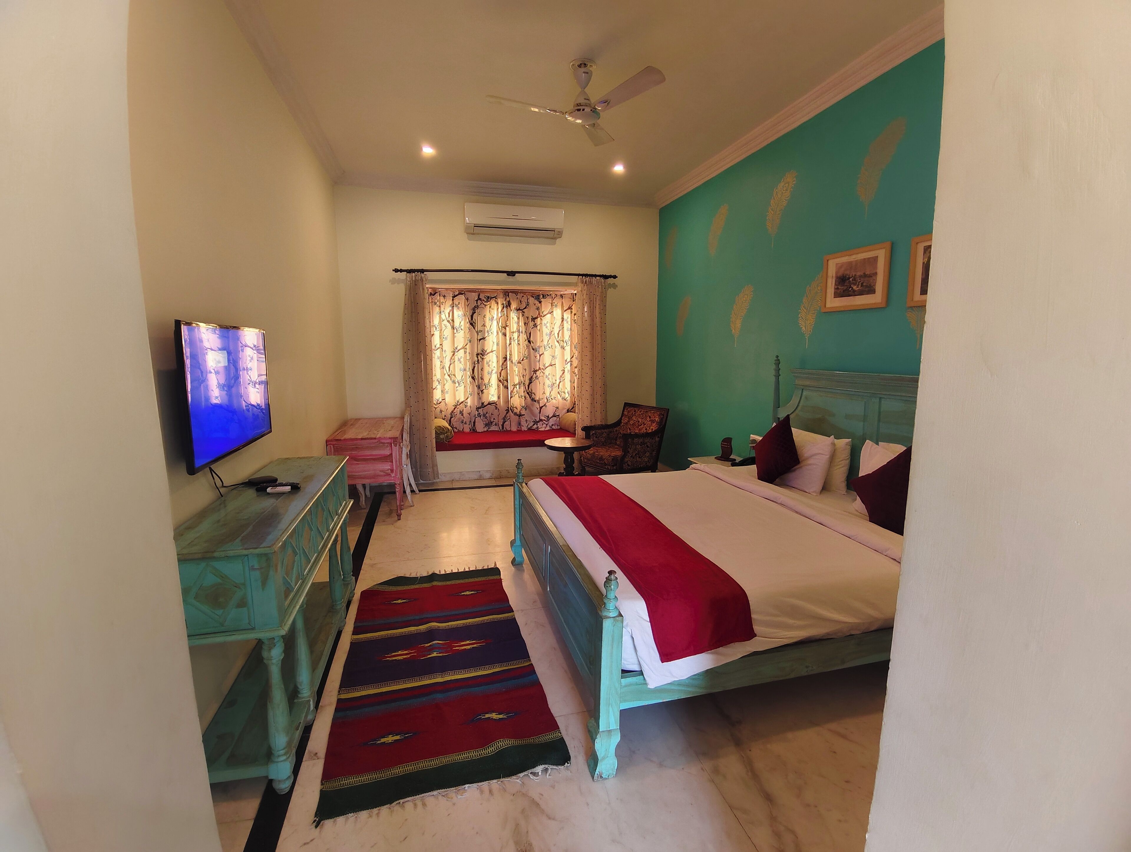A bedroom with a bed, a television, a ceiling fan, a window with curtains, and a wall with feather patterns.