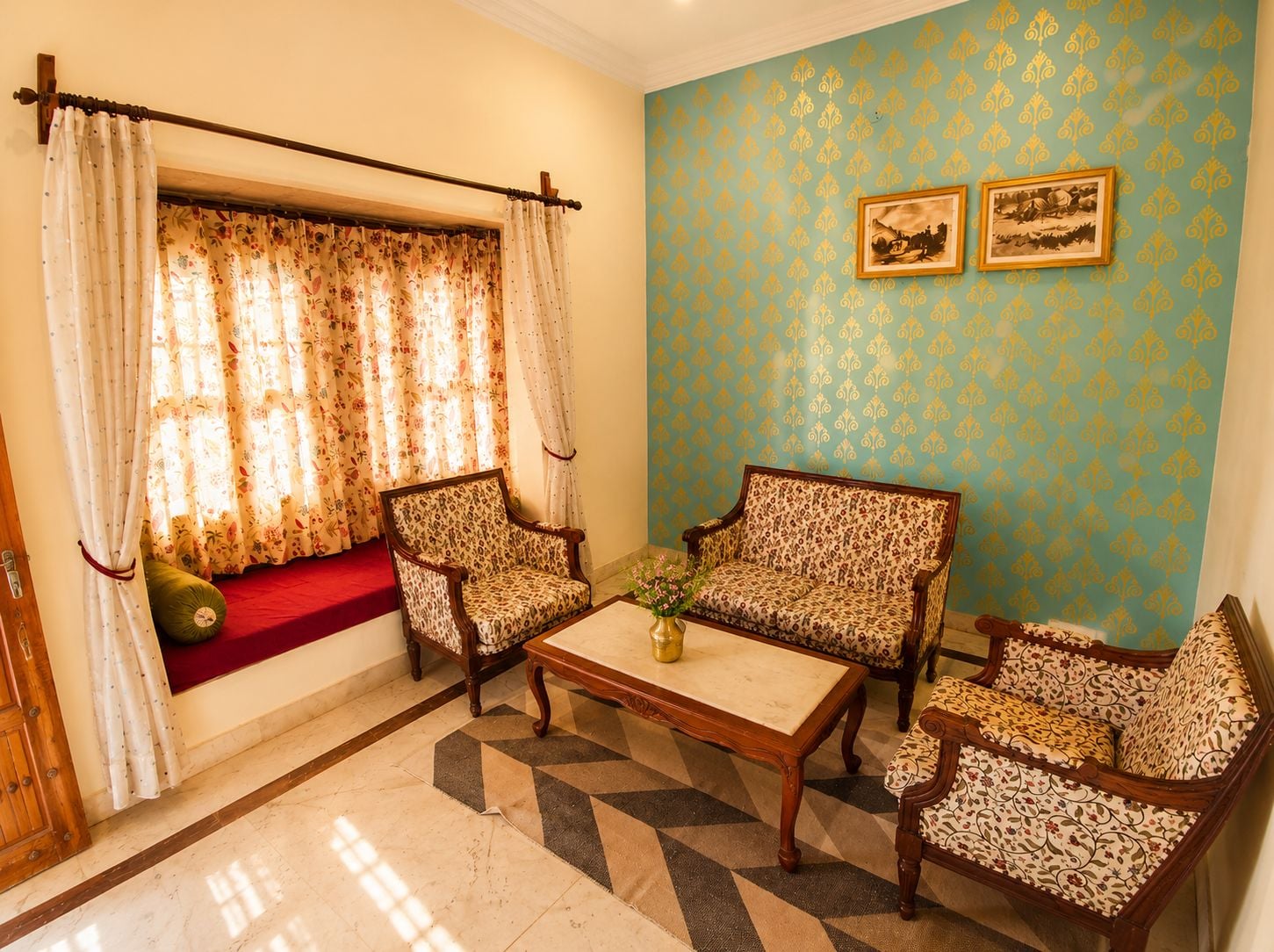 Una habitación con dos sillones estampados, una mesa de centro, un sofá rojo y una ventana con cortinas florales.