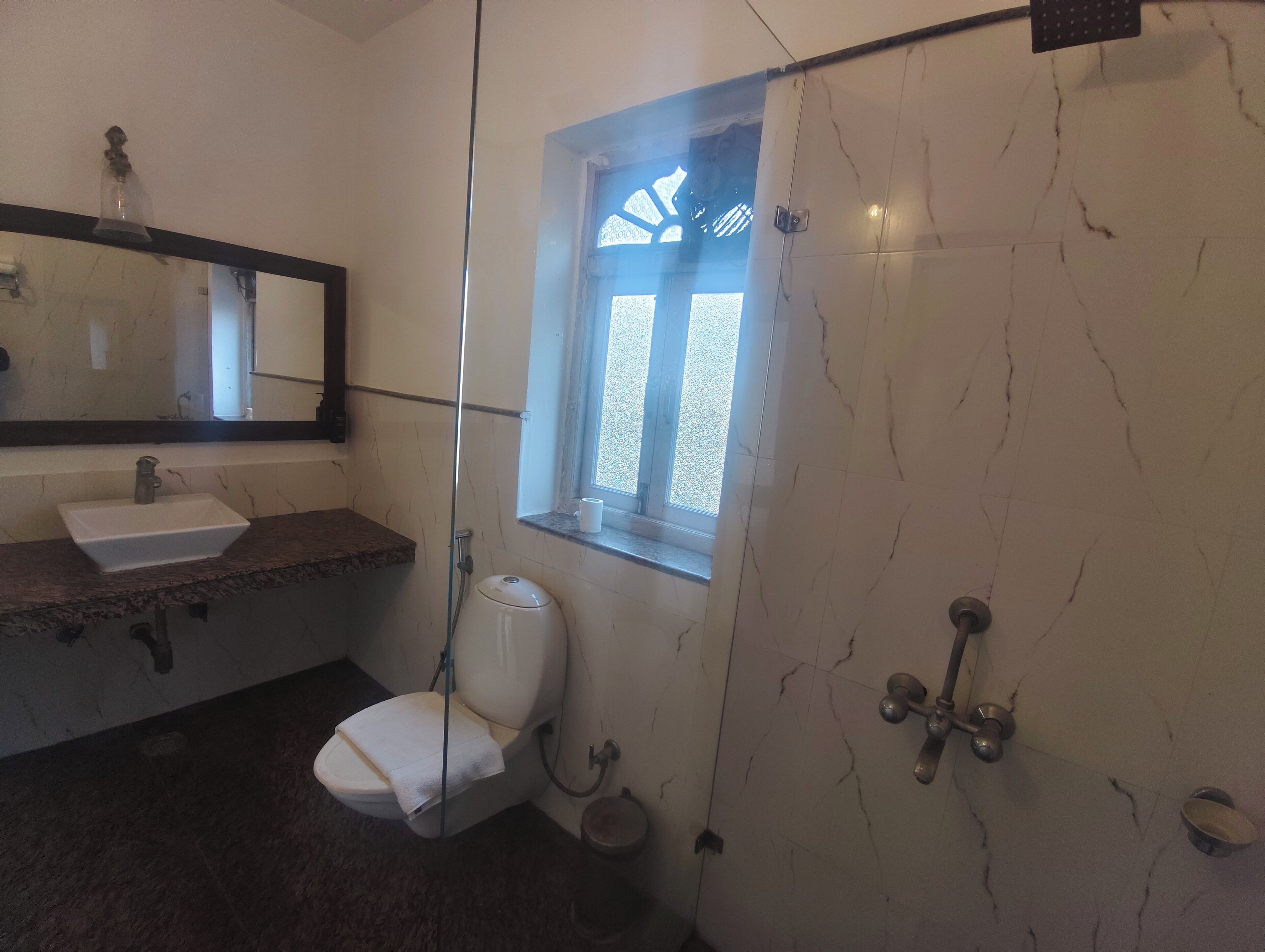 Une salle de bain avec un lavabo blanc, des toilettes, une douche avec une porte en verre et une fenêtre donnant sur l’extérieur.