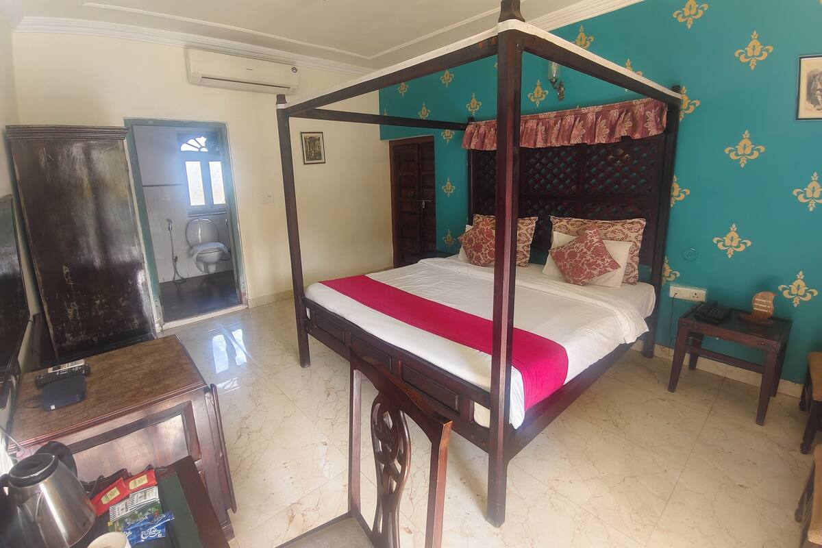 A four-poster bed with a red bedspread, a wooden desk with a kettle and tea bags, and a small bedside table with a lamp.