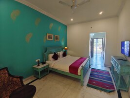 Una habitación con paredes verde azulado, una cama con una colcha roja, una mesita de noche con una lámpara, un televisor y una alfombra estampada.