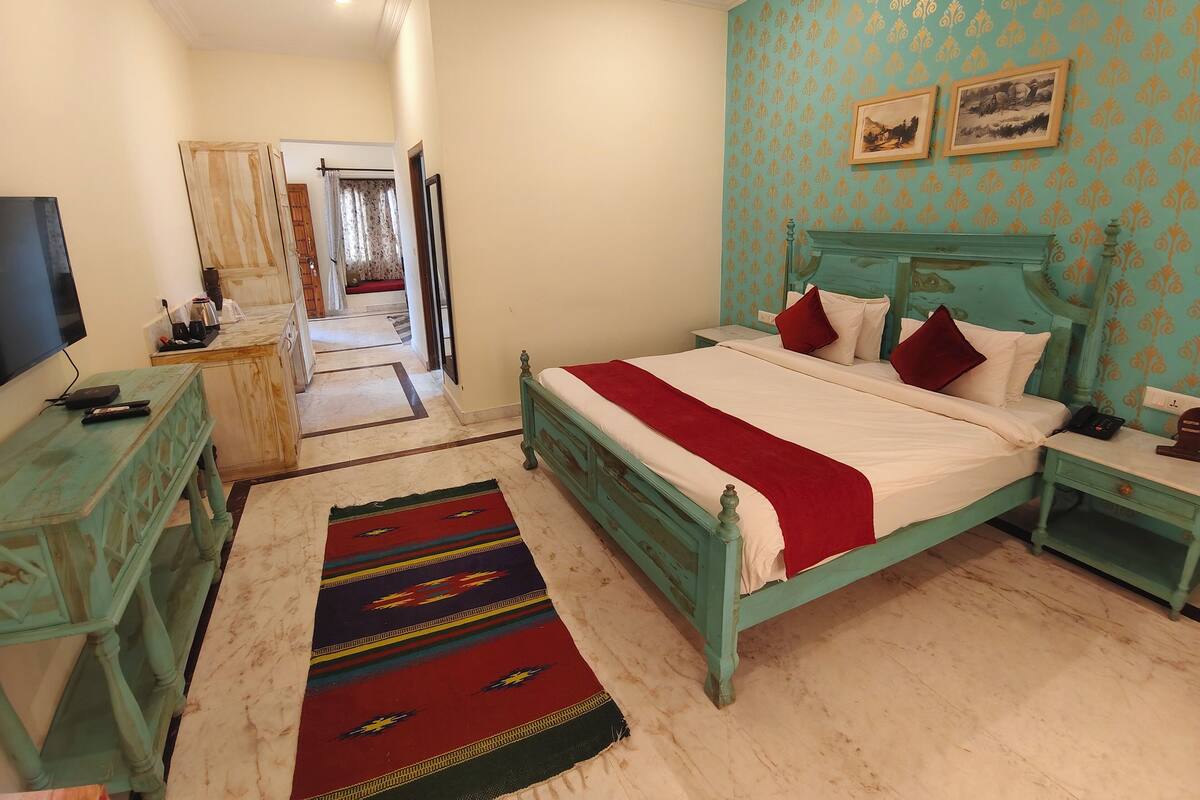 A bedroom with a bed, a TV, a desk, and a door leading to another room.