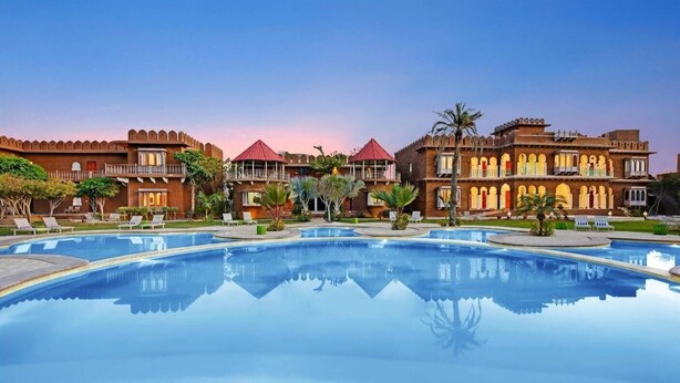 A hotel with a large swimming pool, multiple buildings with traditional architecture, and a clear sky.