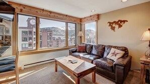 Un salon chaleureux avec un canapé en cuir, une table basse en bois et une grande fenêtre donnant sur des immeubles et des montagnes.
