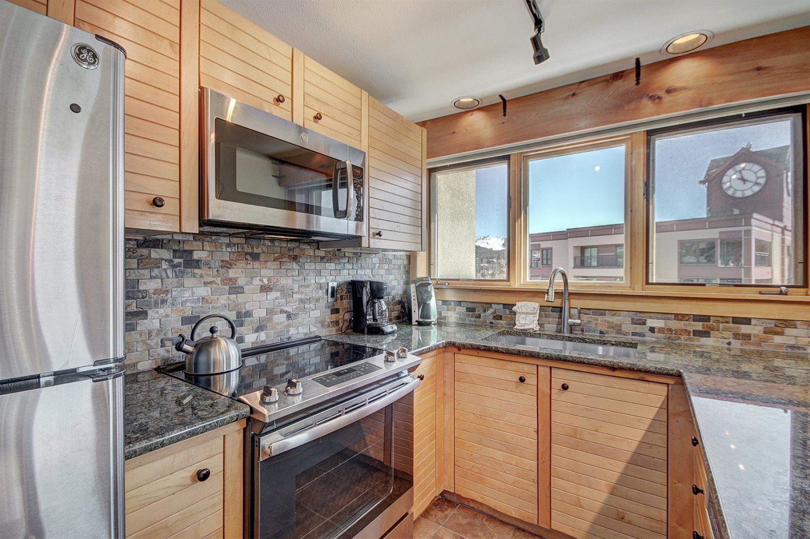 A modern kitchen with wooden cabinets, a stainless steel refrigerator, a built-in microwave, and a large window with a view of a clock tower.