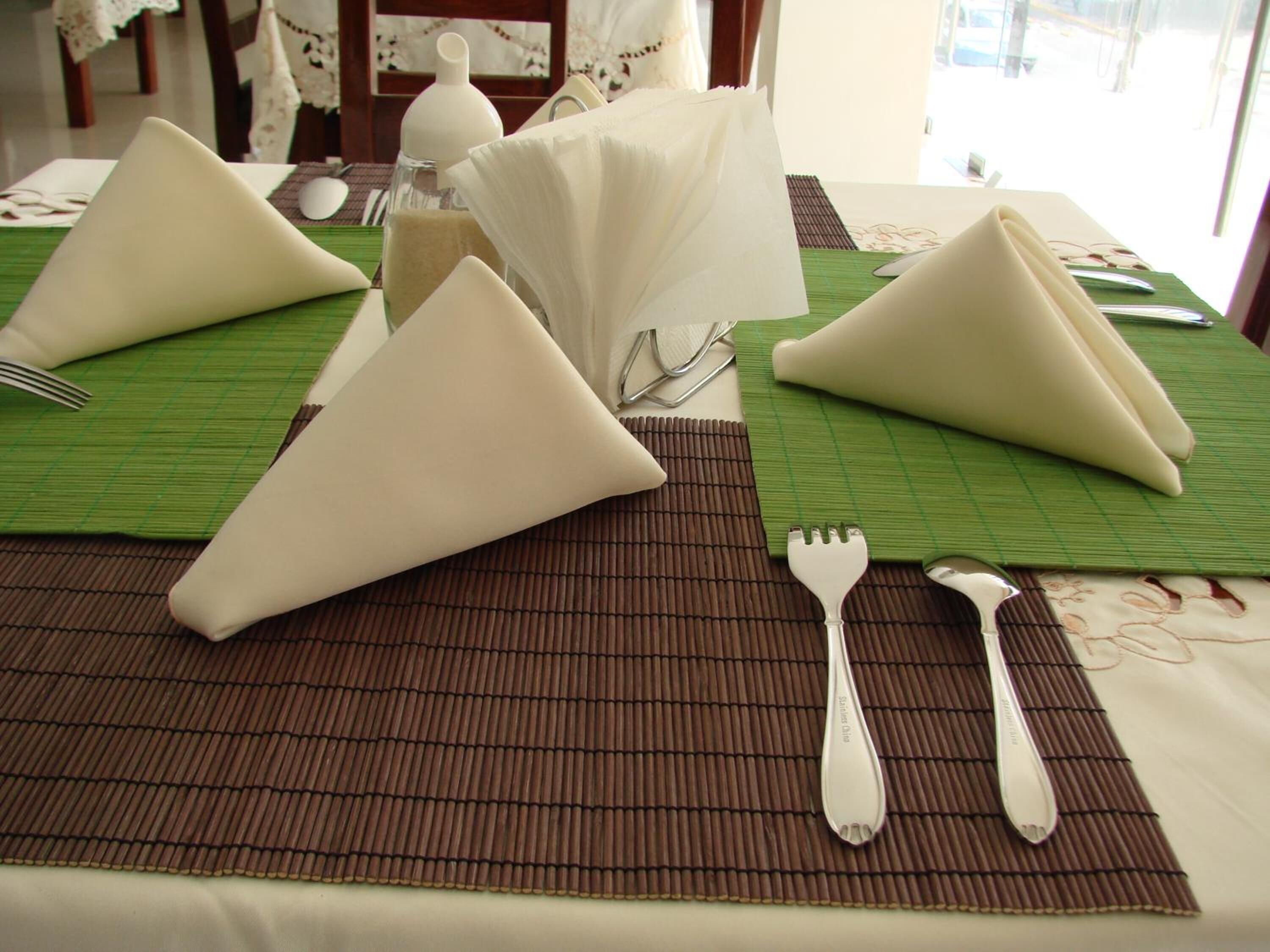Una mesa con servilletas blancas dobladas en forma de triángulo, un tenedor y una cuchara sobre un mantel de bambú.