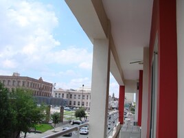 Un balcón con vista a un paisaje urbano, que incluye un edificio grande de arquitectura clásica y una calle concurrida con coches.