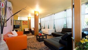 A living room with a sofa, armchairs, a coffee table, and a TV. There is a large window with curtains, a rug, and a plant. - Hotel Milano (Gatteo)