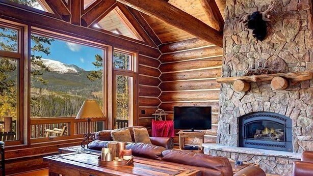 A log cabin interior with a stone fireplace, wooden furniture, and large windows overlooking a mountainous landscape. - Big Timber Lodge (Breckenridge)