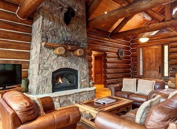 A log cabin interior with a stone fireplace, leather sofas, and a television.