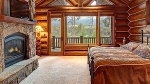 A log cabin bedroom with a stone fireplace, a bed, and a balcony with a view of trees. - Big Timber Lodge (Breckenridge)