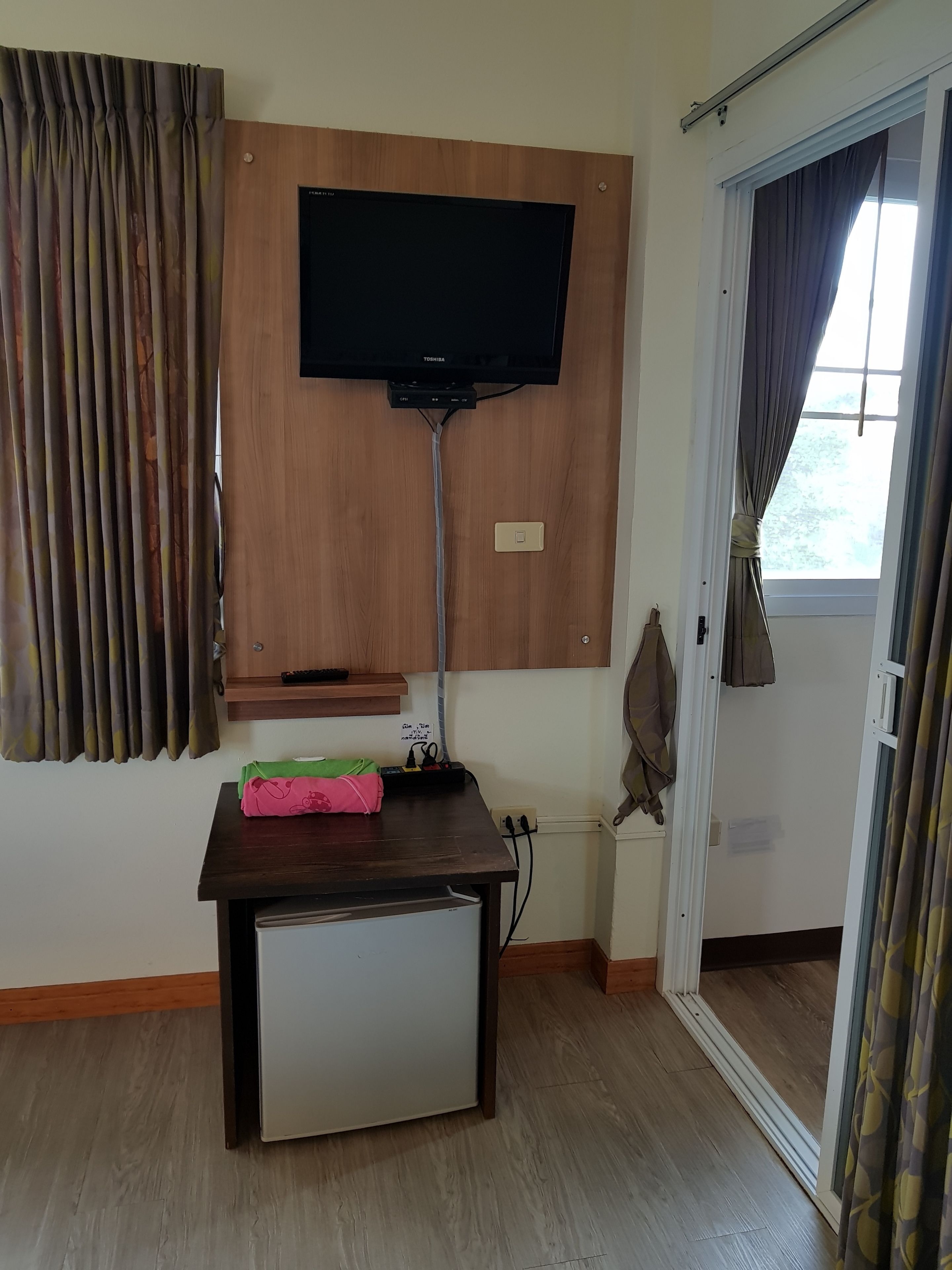 A room with a mounted TV, a small refrigerator, a power strip, and a door leading to a balcony.