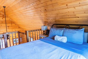 A wooden cabin bedroom with a bed, pillows, a bedside table with a lamp, and a view of the outdoors. - Holmstead Ranch Resort (Veyo)