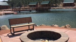 A wooden bench and a stone fire pit on a concrete deck by a body of water, with cabins in the background. - Holmstead Ranch Resort (Veyo)