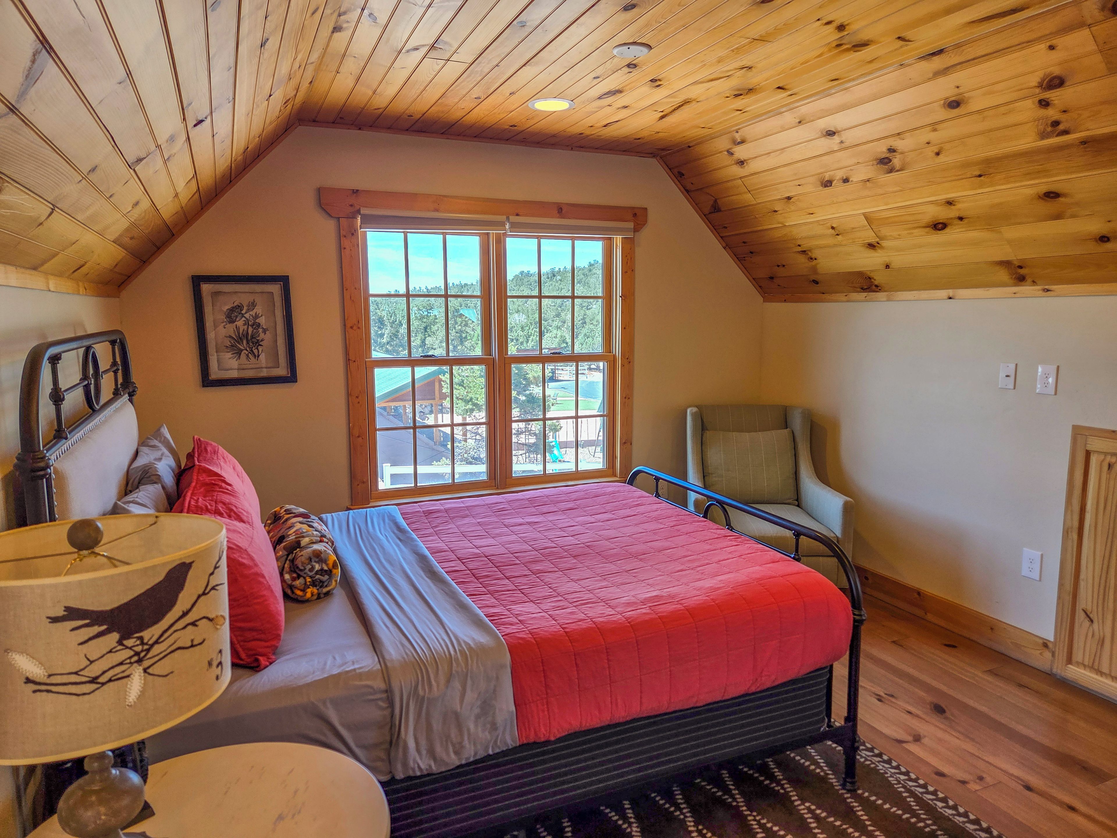 A bedroom with a wooden ceiling, a large window, a bed with a red blanket, a chair, and a lamp.