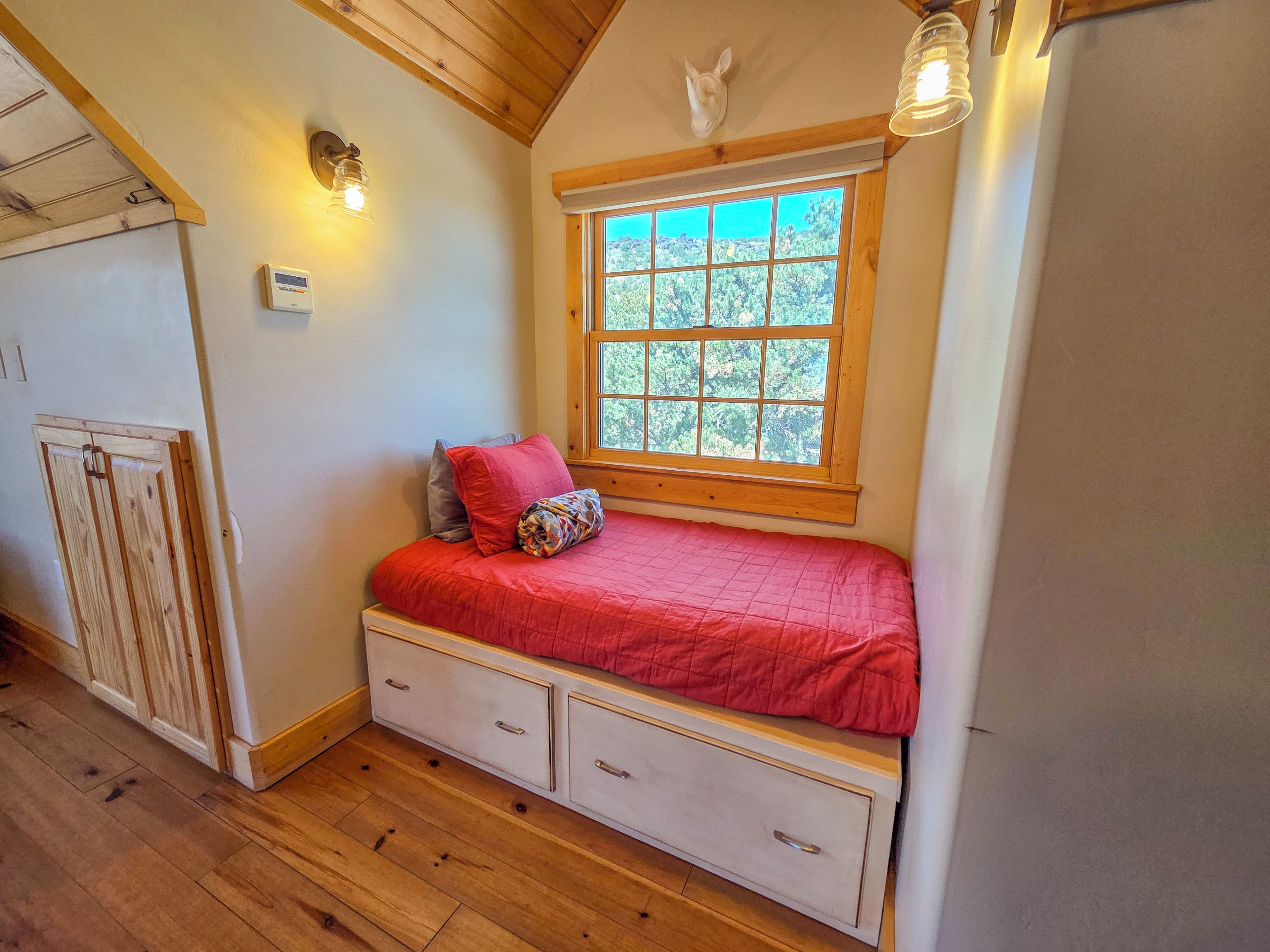 A bedroom with a wooden ceiling, a window with a view, a bed with a red blanket, and a small dresser.