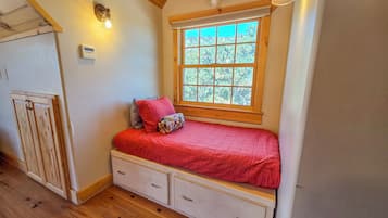 A bedroom with a wooden ceiling, a window with a view, a bed with a red blanket, and a small dresser.