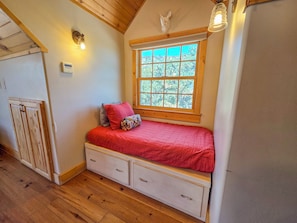 A bedroom with a wooden ceiling, a window with a view, a bed with a red blanket, and a small dresser.