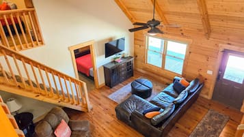 Een loft-achtige kamer met houten plafond, een trap, een zithoek, een televisie en een raam met uitzicht op een terras.