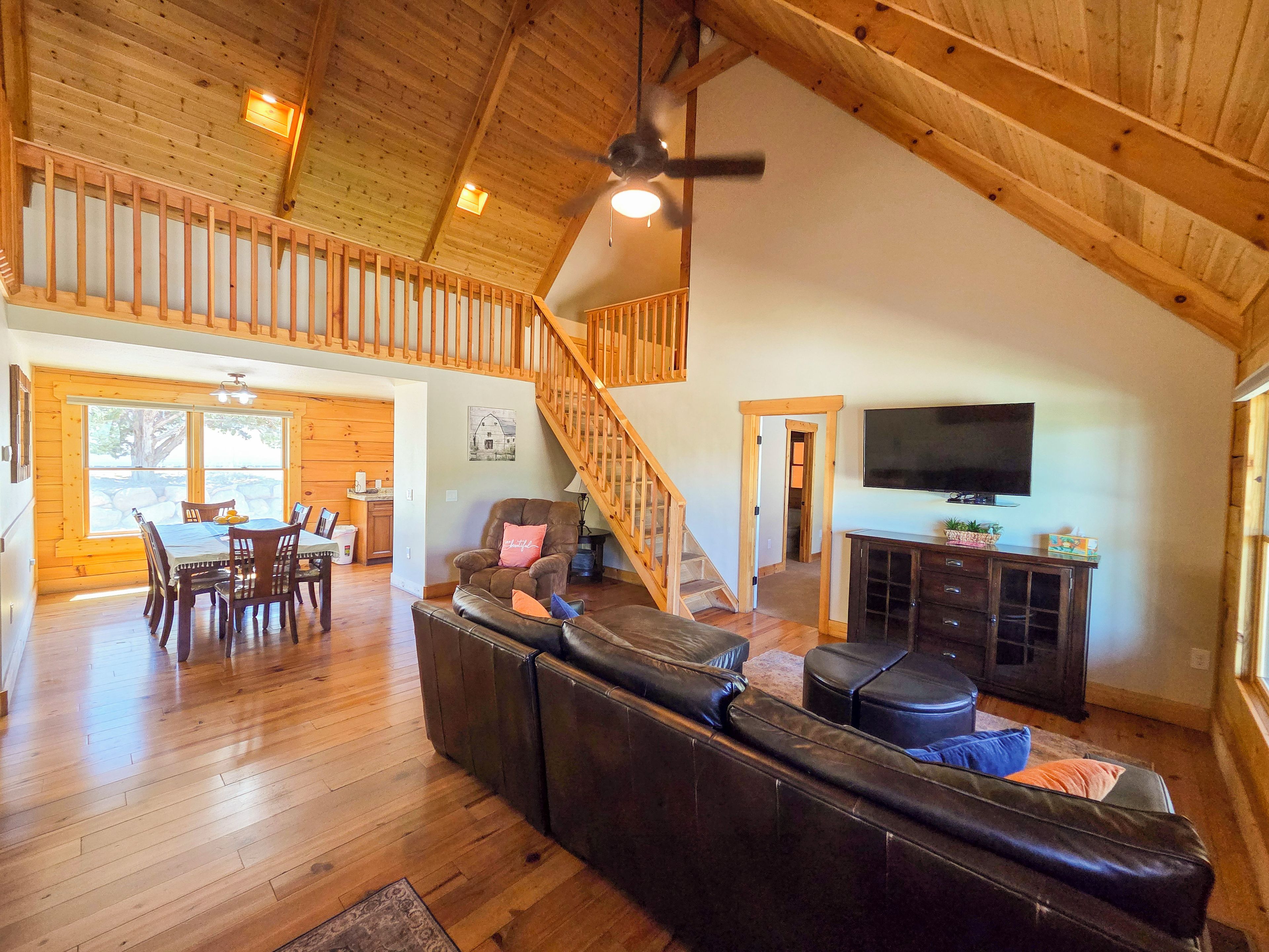 A spacious living area with a high ceiling, wooden beams, a dining table with chairs, a leather sofa, a television, and a staircase.