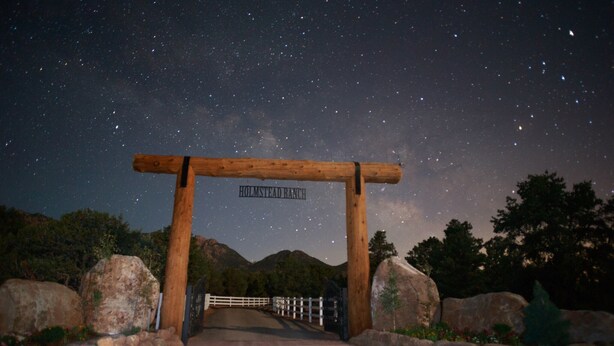 A wooden archway with "HOLMSTEAD RANCH" signage, leading to a road with a white fence and large rocks on either side. - Holmstead Ranch Resort (Veyo)