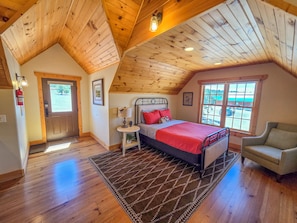 A bedroom with a sloped ceiling, a bed with a red bedspread, a chair, a small table with a lamp, and a window with a view of the outside. - Holmstead Ranch Resort (Veyo)
