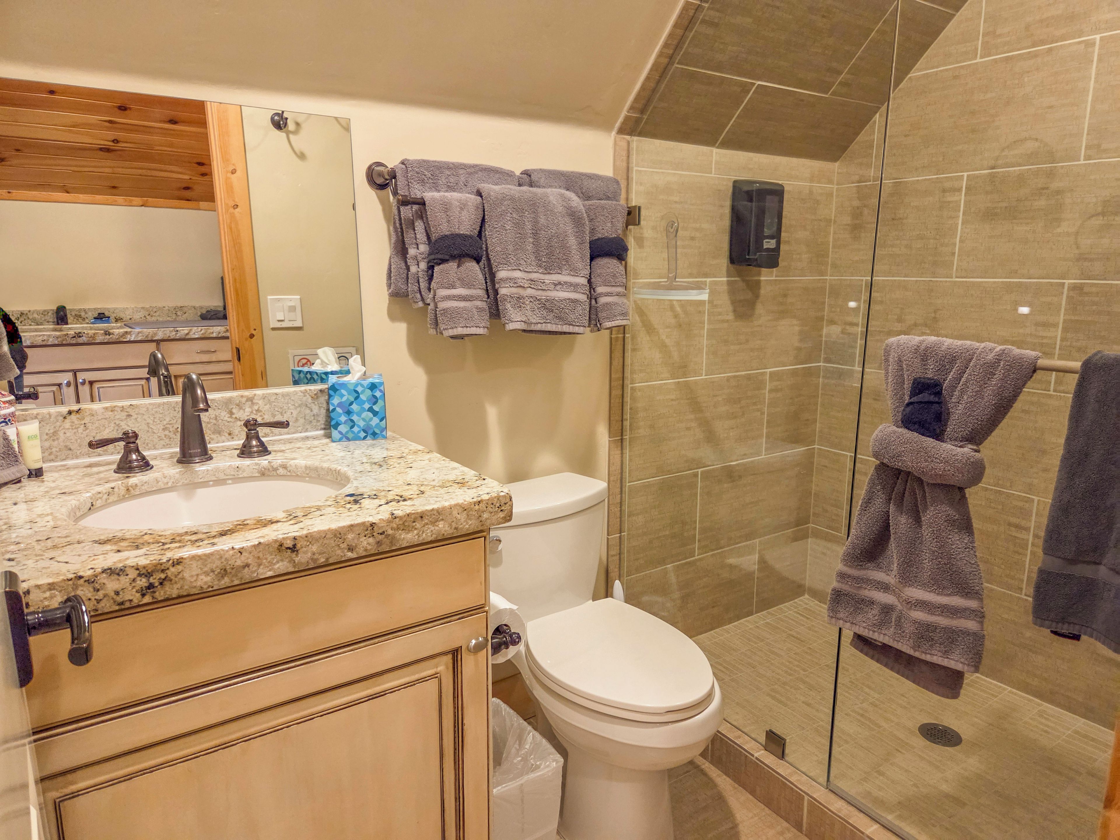 A bathroom with a granite countertop, a toilet, a glass shower enclosure, and a towel rack with multiple towels.