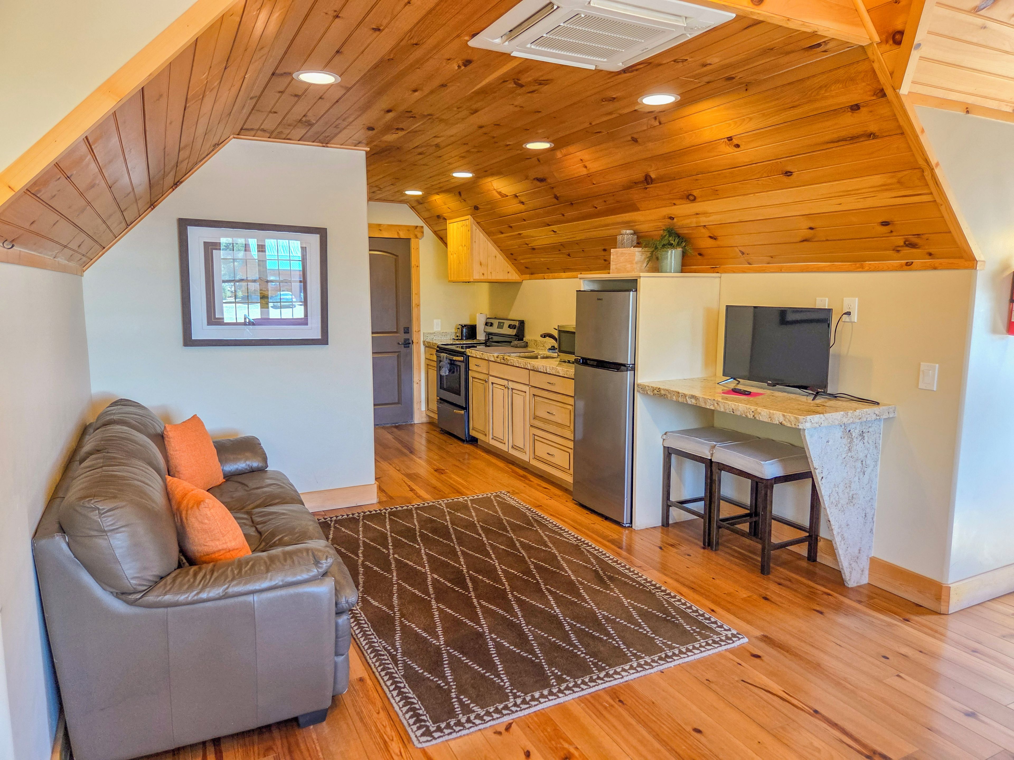A cozy living room with a gray sofa and a patterned rug, a kitchen with wooden cabinets and a stainless steel refrigerator, and a dining area with a small table and chairs.