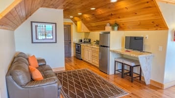 A cozy living room with a gray sofa and a patterned rug, a kitchen with wooden cabinets and a stainless steel refrigerator, and a dining area with a small table and chairs.