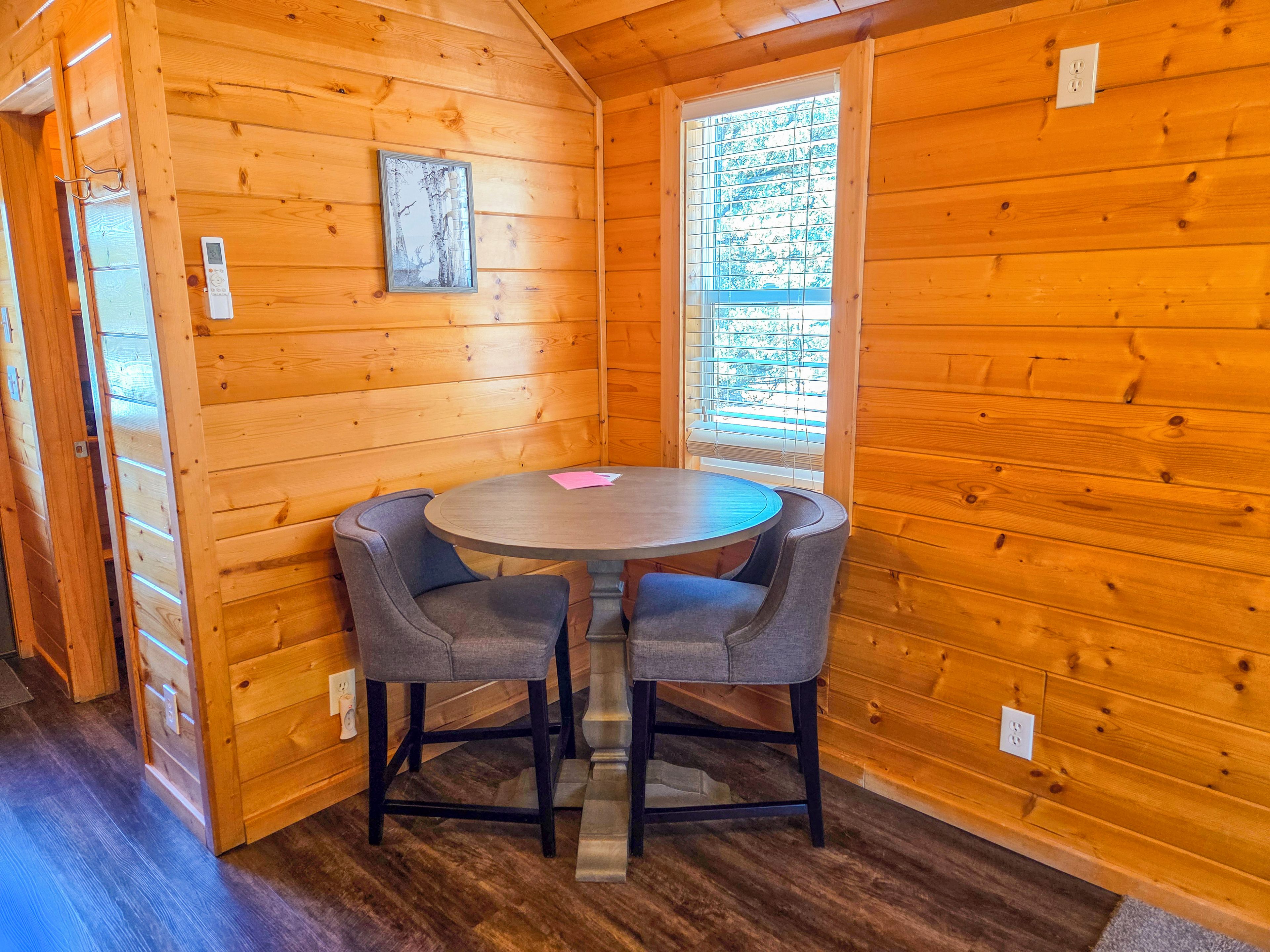 A wooden cabin interior with a round table and two chairs, a window with blinds, and a framed picture on the wall.