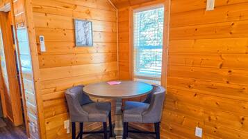 A wooden cabin interior with a round table and two chairs, a window with blinds, and a framed picture on the wall.