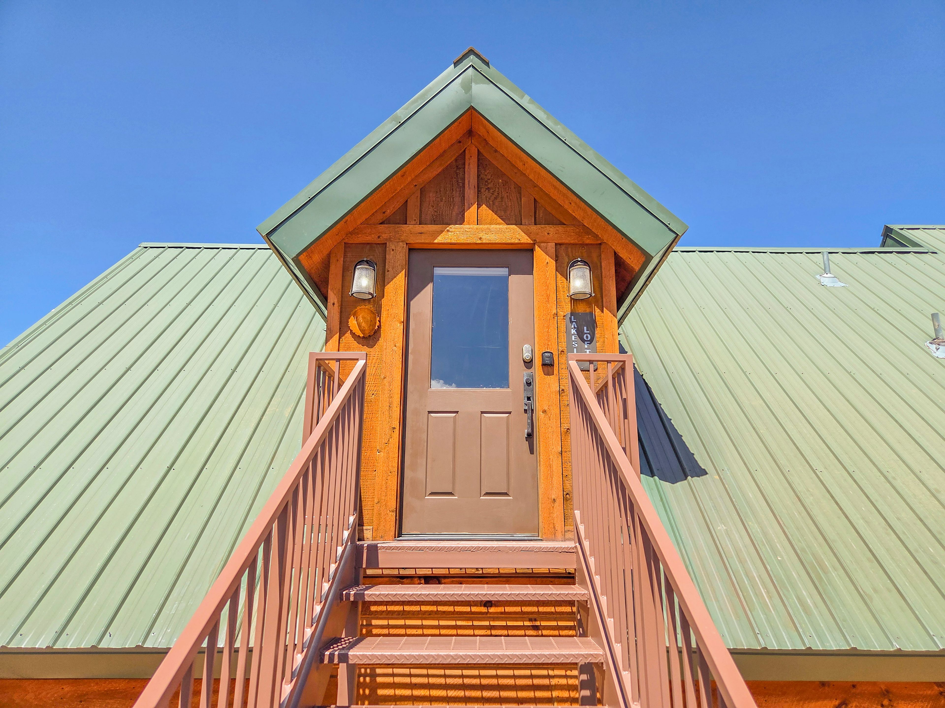 Une cabane en bois avec un toit en métal vert, une porte centrale surmontée d’un petit porche et une lanterne fixée au mur.