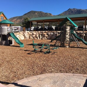 A playground with a slide and climbing structures, surrounded by a stone wall and a covered seating area. - Holmstead Ranch Resort (Veyo)