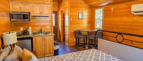 A log cabin with a kitchenette, a dining area, and a bed with a patterned bedspread.