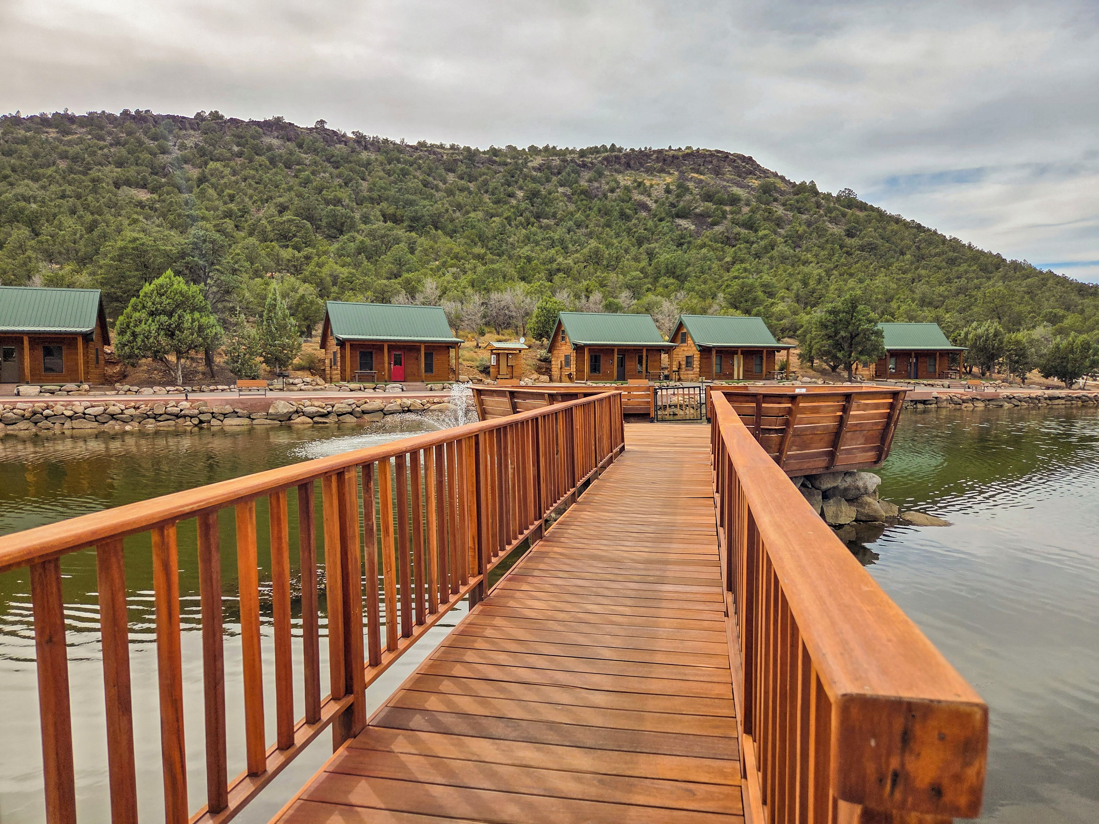 A wooden bridge leading to cabins with green roofs, surrounded by a forested hillside.