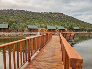 A wooden bridge leading to cabins with green roofs, surrounded by a forested hillside. - Holmstead Ranch Resort (Veyo)